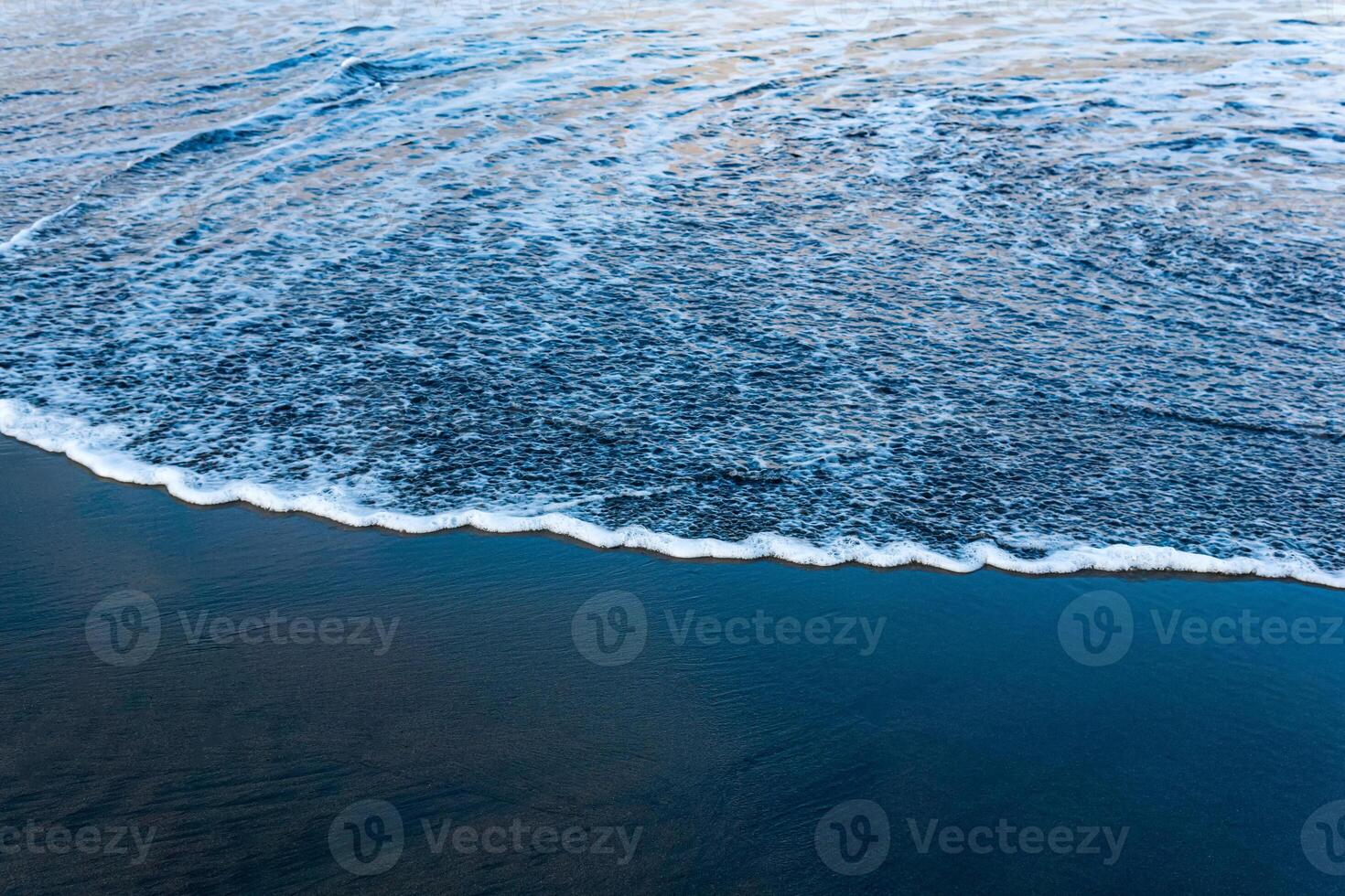 oceaan strand met zwart vulkanisch zand, de lucht is weerspiegeld in de gerold terug Golf van de surfen foto