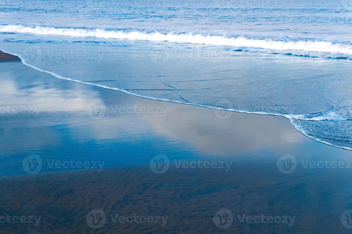 oceaan strand met zwart vulkanisch zand, de lucht is weerspiegeld in de gerold terug Golf van de surfen foto