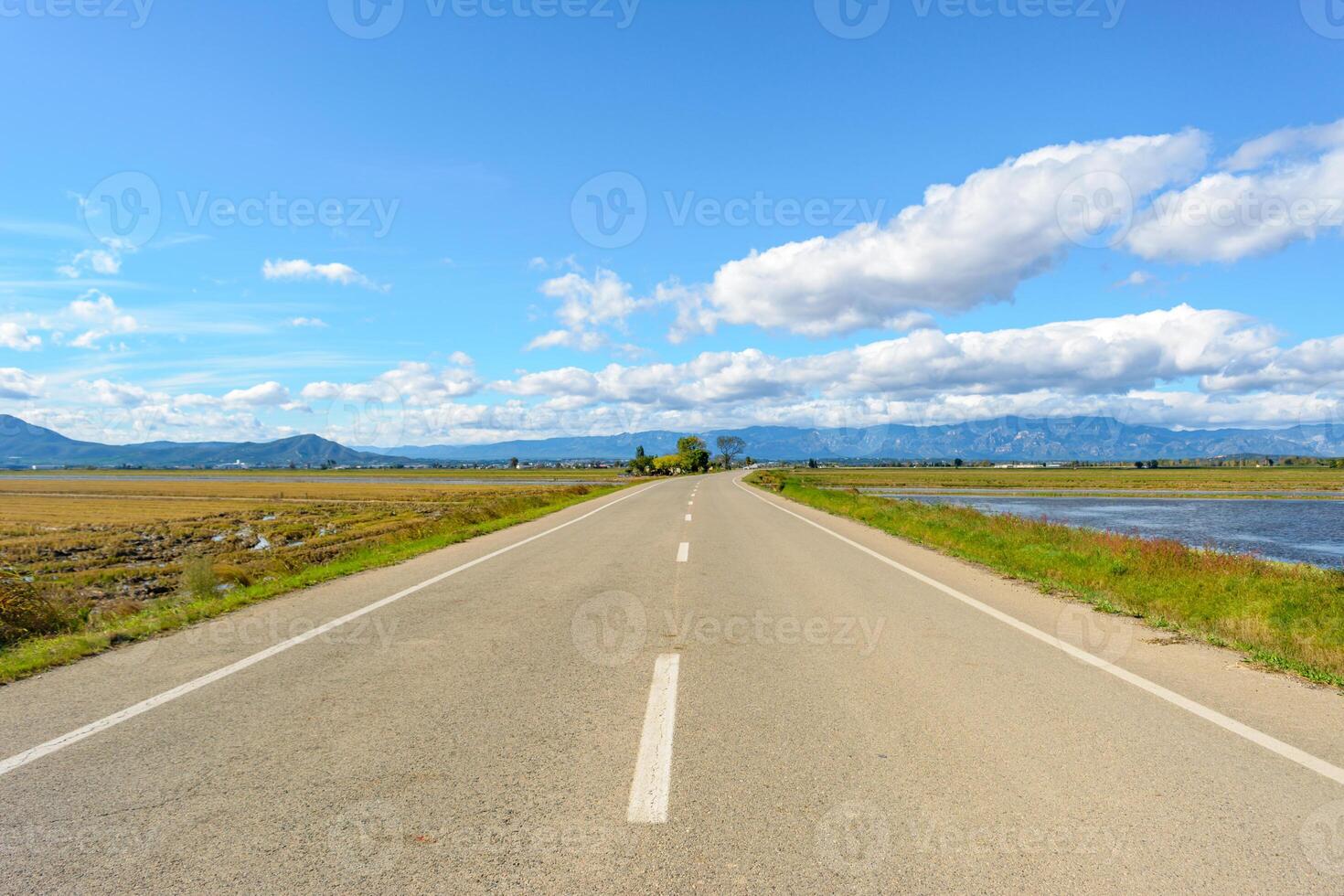 een Rechtdoor weg leidend naar bergen onder een helder blauw lucht met wolken, visie van eenzaam weg in de ebro delta, tarragona, Catalonië, Spanje foto