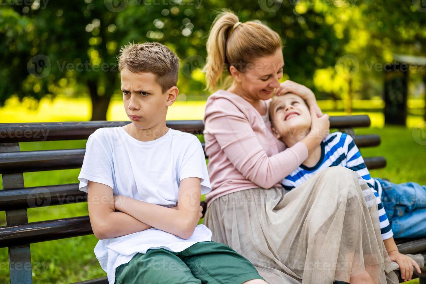 gelukkig moeder is zittend met haar zonen Aan bank in park. een jongen is beledigd en pruilend foto