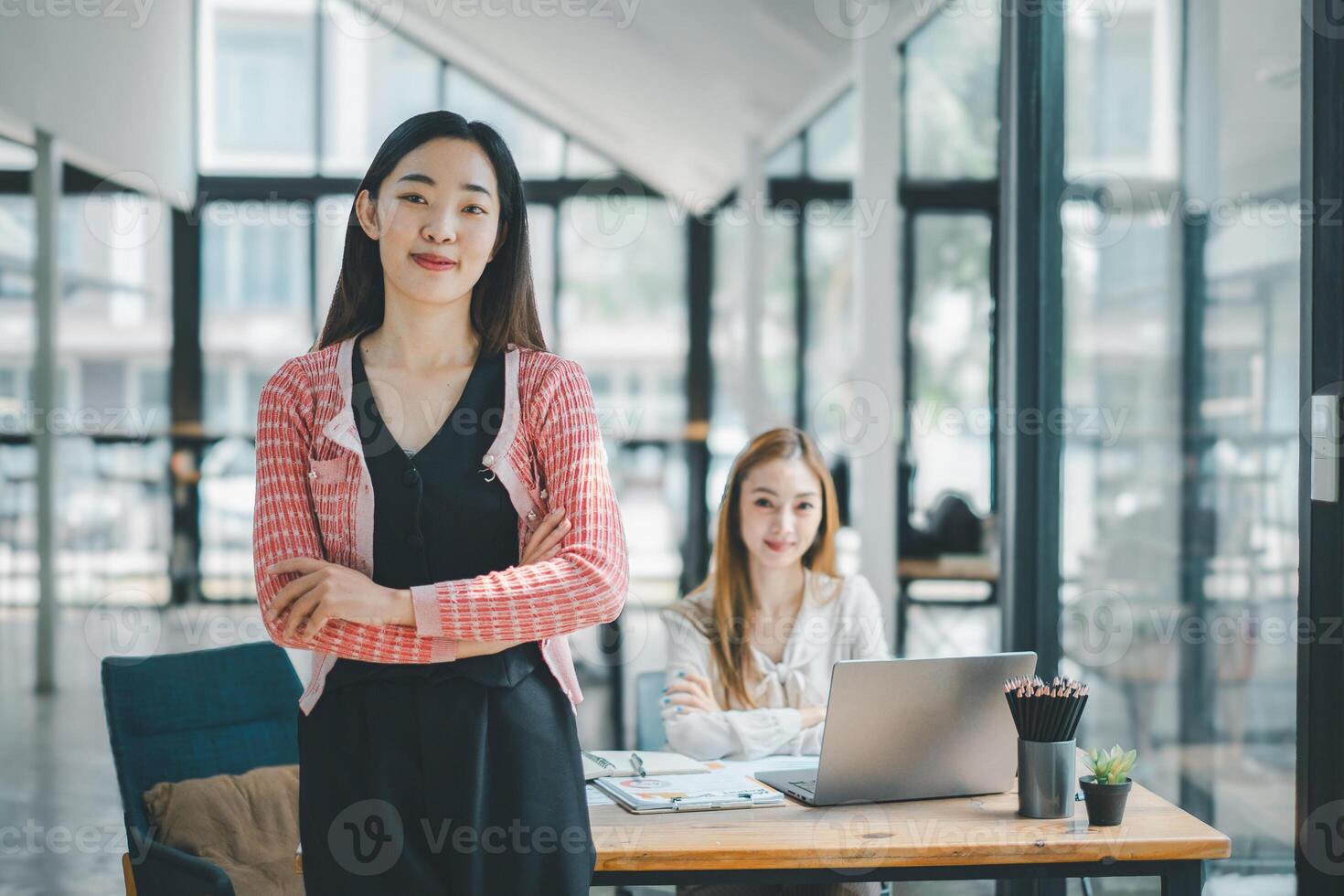 bedrijf team analytics concept, een zelfverzekerd jong vrouw staand armen gekruiste met haar collega in de achtergrond in een modern, licht gevuld kantoor omgeving. foto