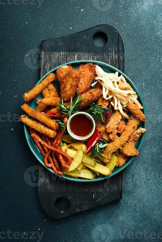 snel voedsel. bord met snacks klompjes, worstjes, Frans Patat, kaas stokken. Aan een zwart steen achtergrond. vrij ruimte voor tekst. foto