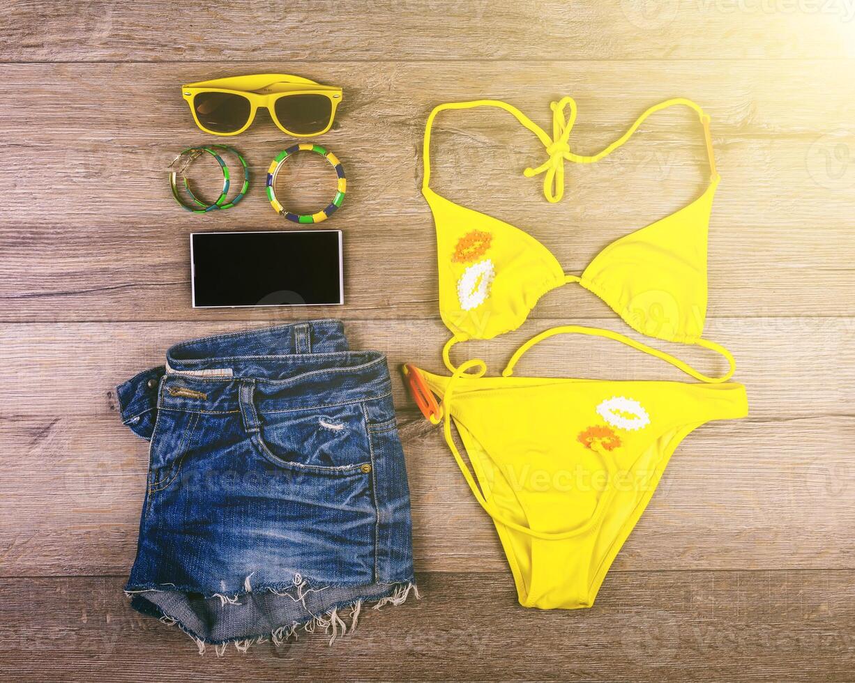 reeks van strand kleren geel bikini, armbanden, jeans korte broek, bril Aan donker houten achtergrond. top visie. zomer vakantie concept. foto