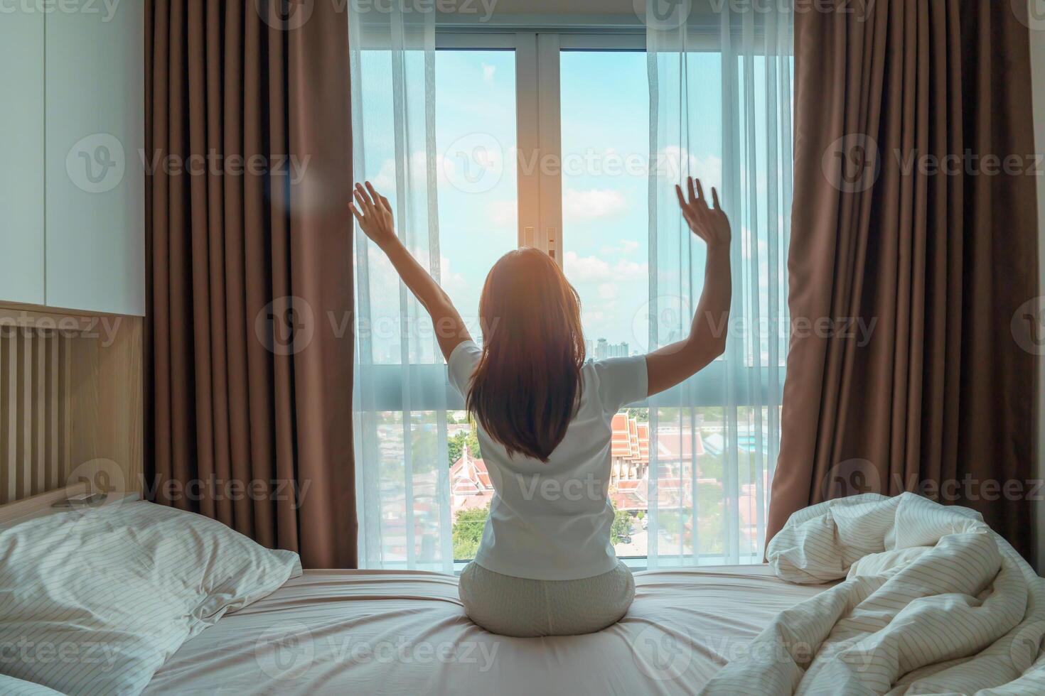 gelukkig vrouw uitrekken Aan bed na wakker worden omhoog, jong volwassen vrouw stijgende lijn armen en op zoek naar venster in de vroeg ochtend. vers kom tot rust en hebben een mooi hoor dag concepten foto