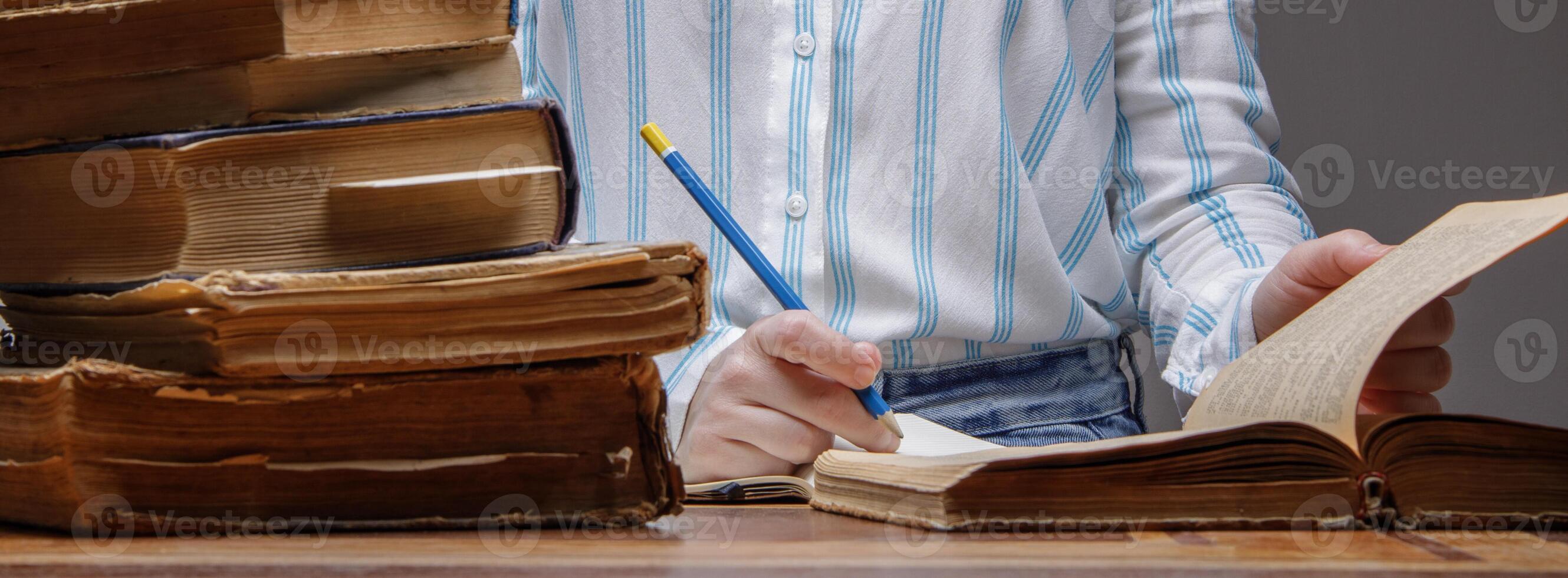 de hand- van een onzichtbaar persoon schrijft met een potlood in een notebook. meerdere oud boeken liggen dichtbij Aan een houten tafel in een Universiteit of school- bibliotheek. wijnoogst papier textuur. selectief focus foto