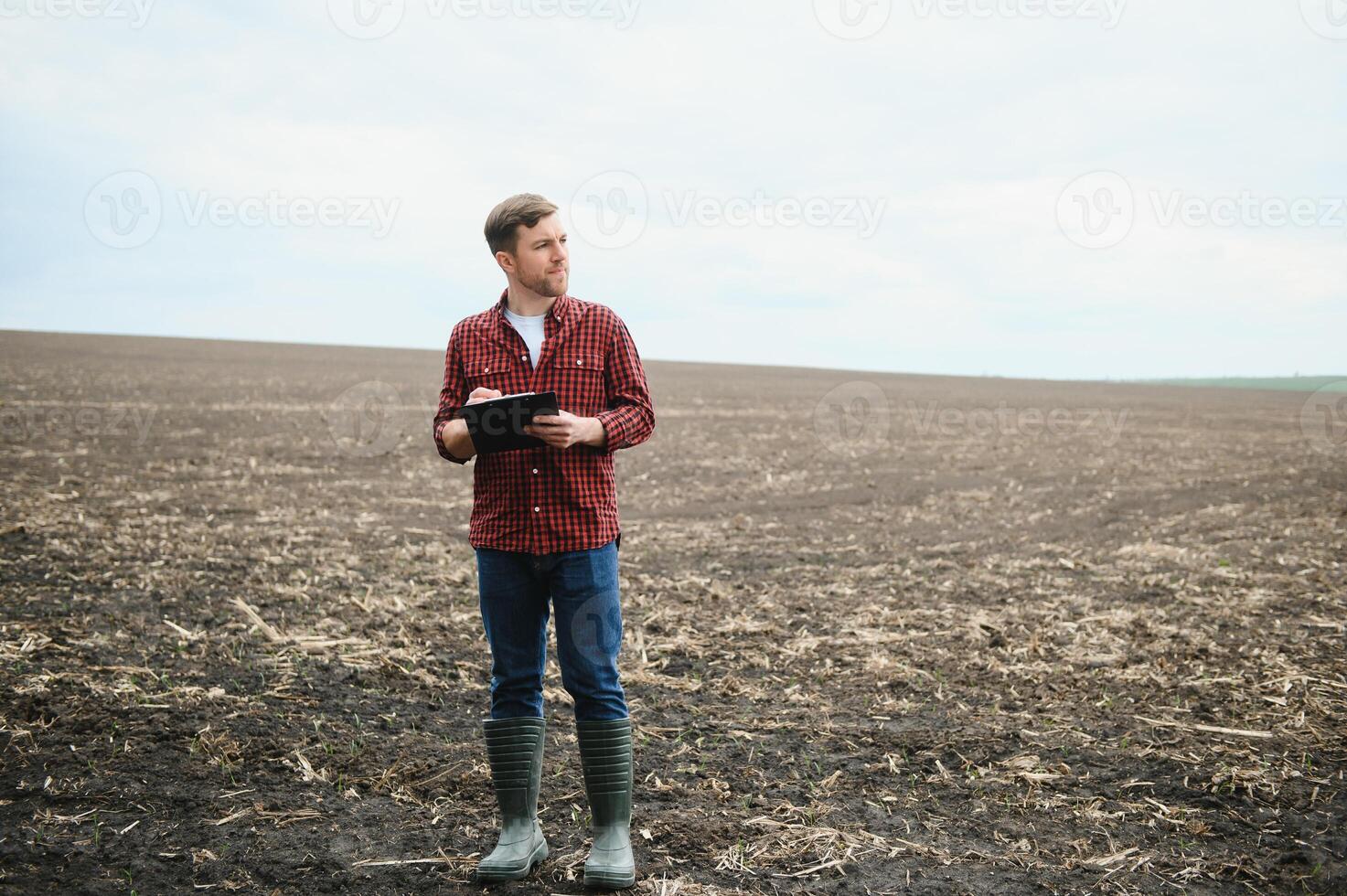 een boer in laarzen werken met zijn tablet in een veld- gezaaid in de lente. een agronoom wandelingen de aarde, beoordelen een geploegd veld- in herfst. landbouw. slim landbouw technologieën foto