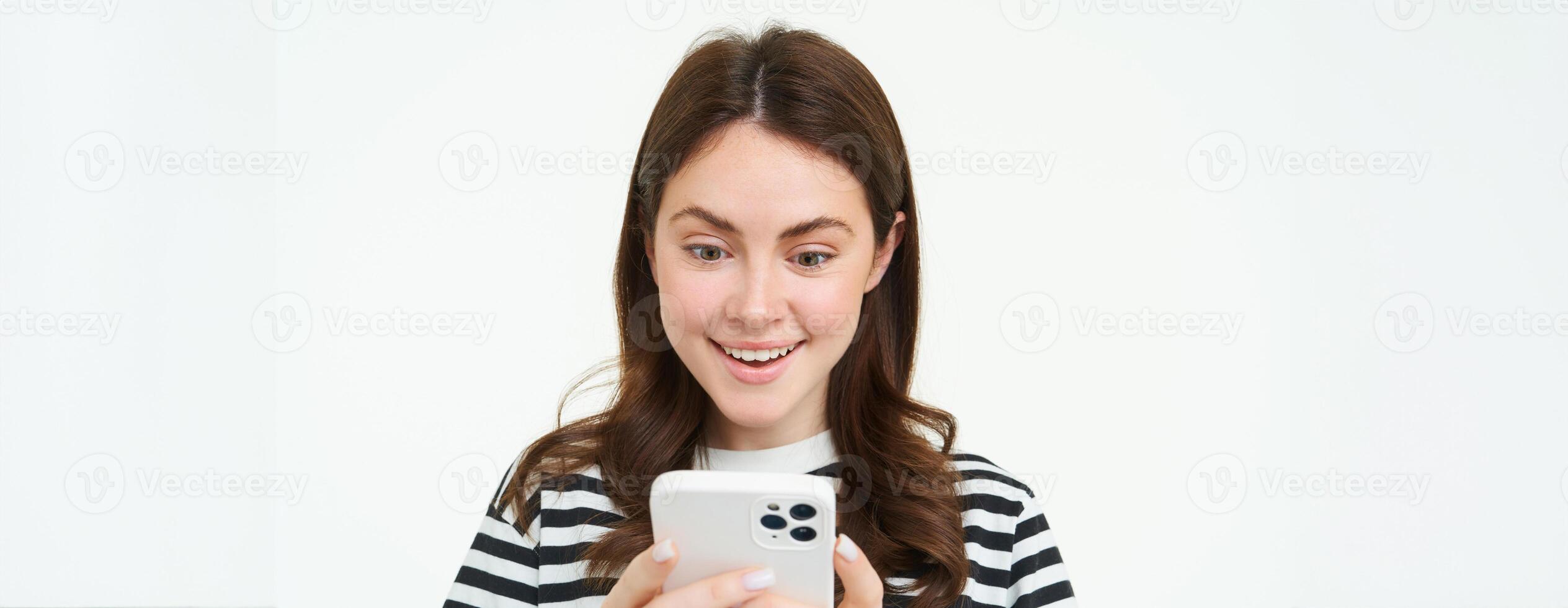 portret van vrouw op zoek verrast Bij mobiel telefoon scherm, leest verbazingwekkend promo in online op te slaan, staand over- wit achtergrond foto