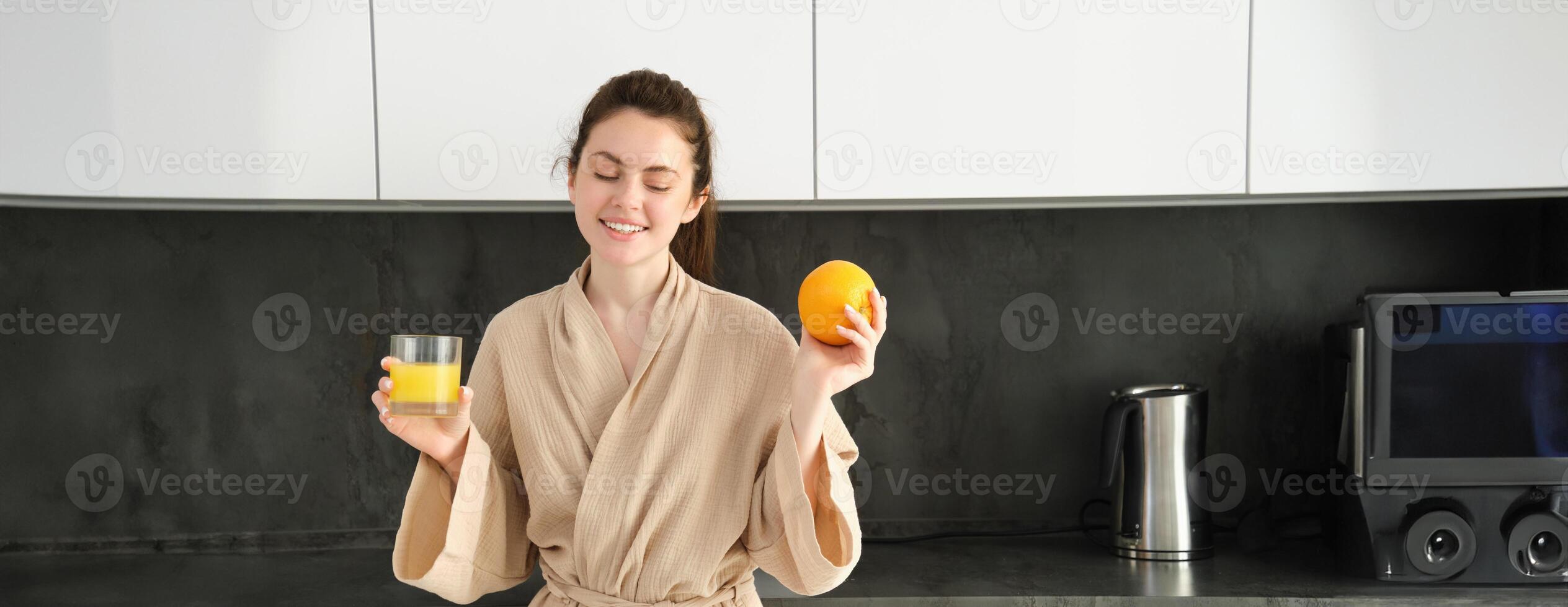 beeld van mooi gezond vrouw in badjas, drinken vers sap, tonen oranje fruit, poseren in keuken foto