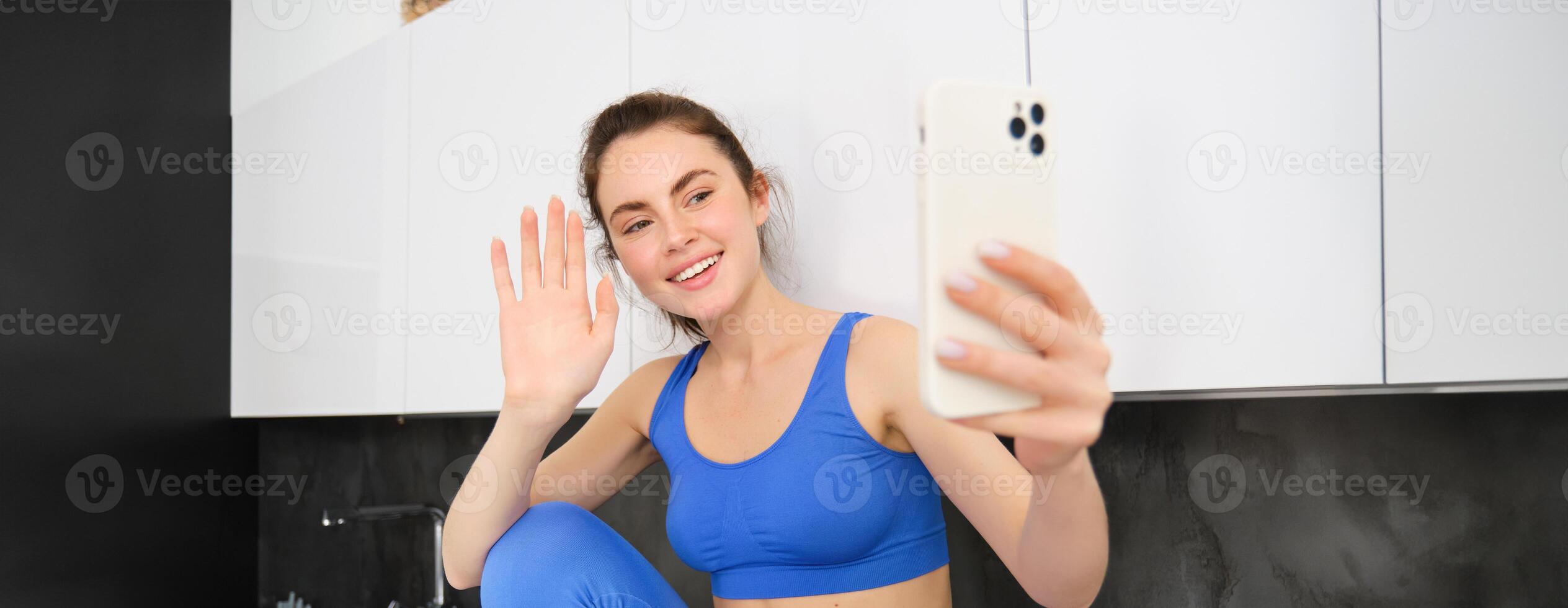 portret van sportvrouw, geschiktheid blogger, opname video, golvend hand- Bij smartphone scherm en lachend, online chatten, zittend in keuken, vervelend activewear foto