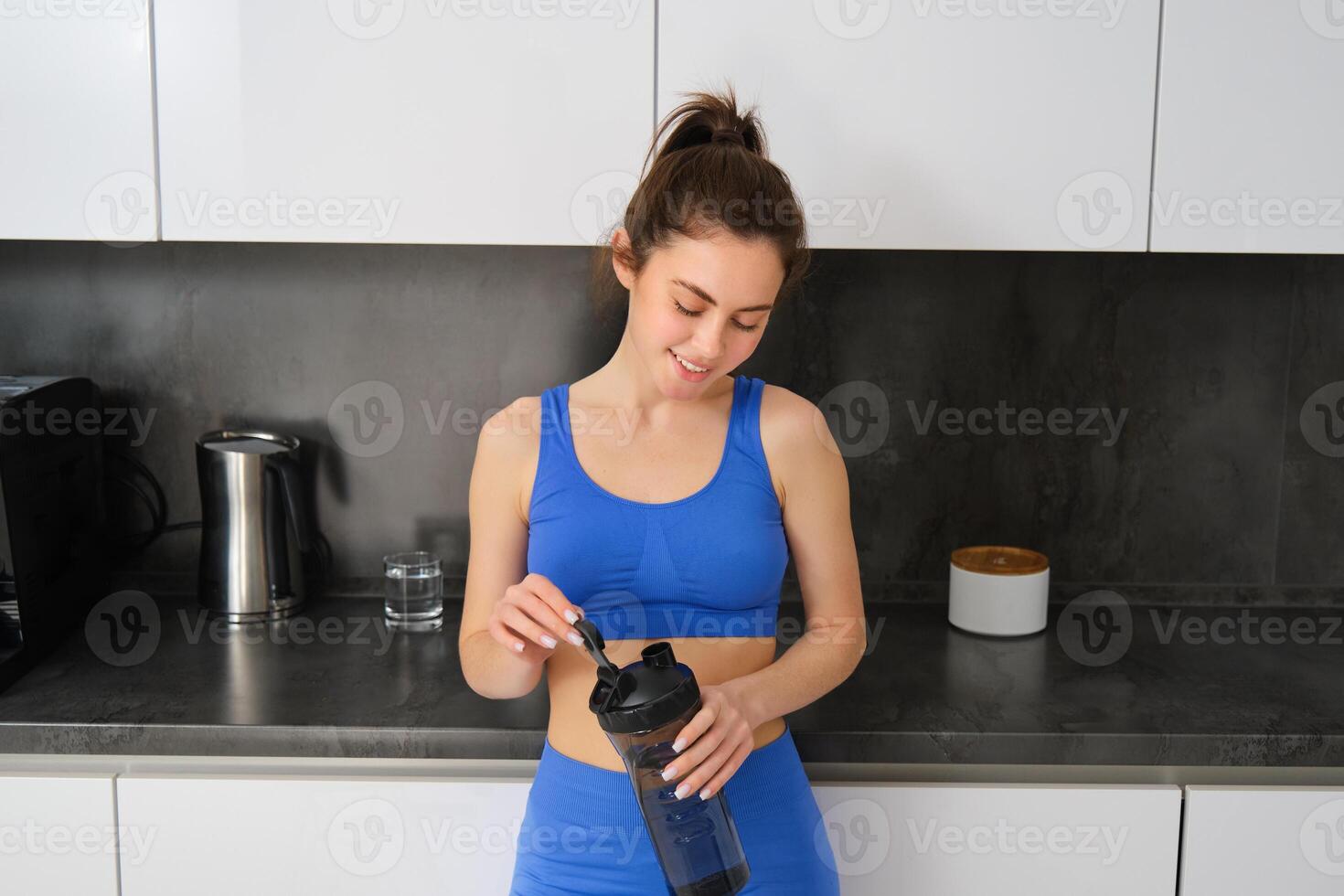 beeld van jong vrouw voorbereidingen treffen dieet supplement, eiwit in haar shaker fles, vervelend geschiktheid kleding, staand in keuken foto