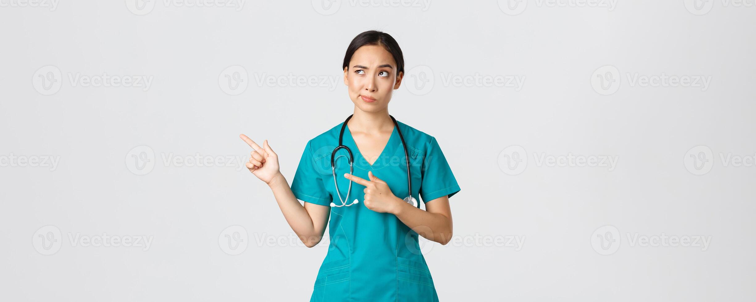 covid19, gezondheidszorg arbeiders, pandemisch concept. sceptisch en twijfelachtig mooi Aziatisch dokter, verpleegster in scrubs grijns, richten bovenste links hoek, op zoek met huiverig aarzelend gezicht, wit achtergrond foto