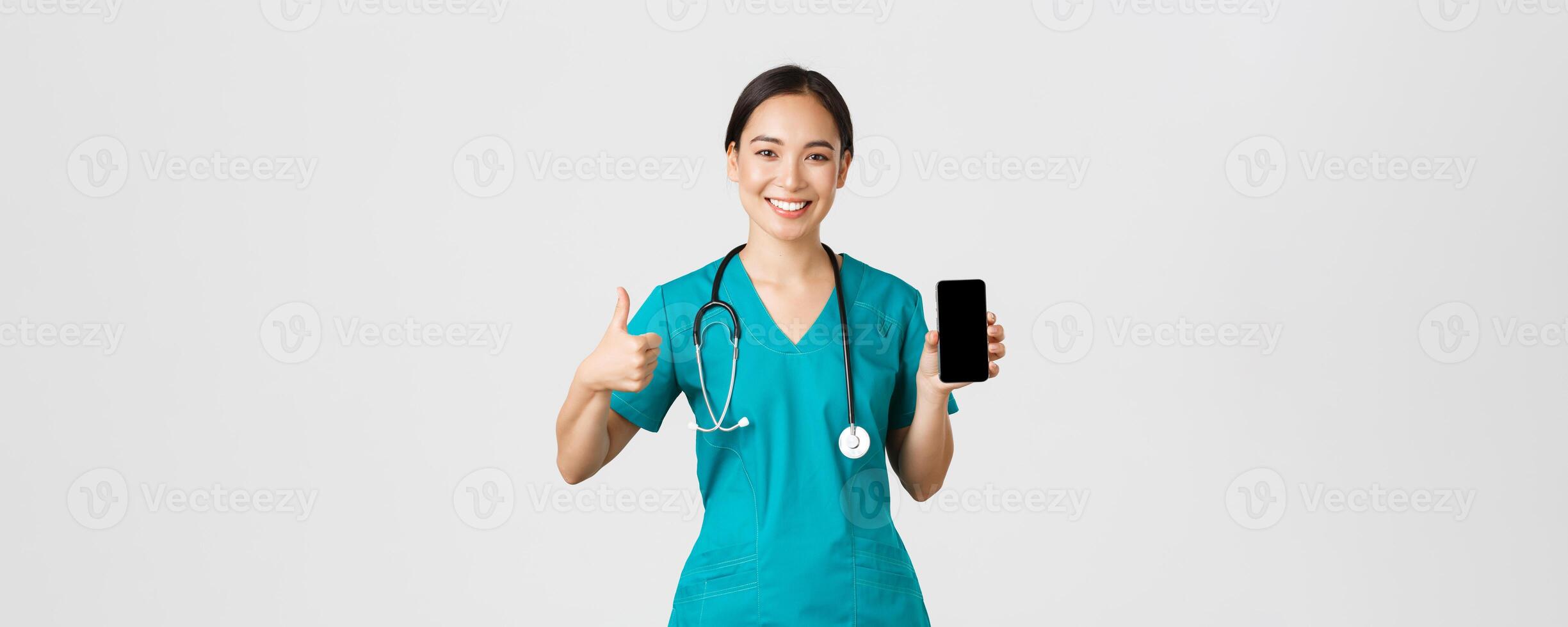 covid19, gezondheidszorg arbeiders en online geneeskunde concept. portret van jong Aziatisch vrouw arts, dokter in scrubs tonen duimen omhoog en smartphone scherm, promoten sollicitatie, wit achtergrond foto