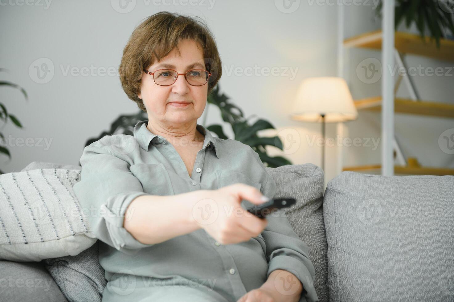 breken nieuws. geschokt senior vrouw aan het kijken TV Bij huis, zittend Aan bankstel met afgelegen controleur in handen foto