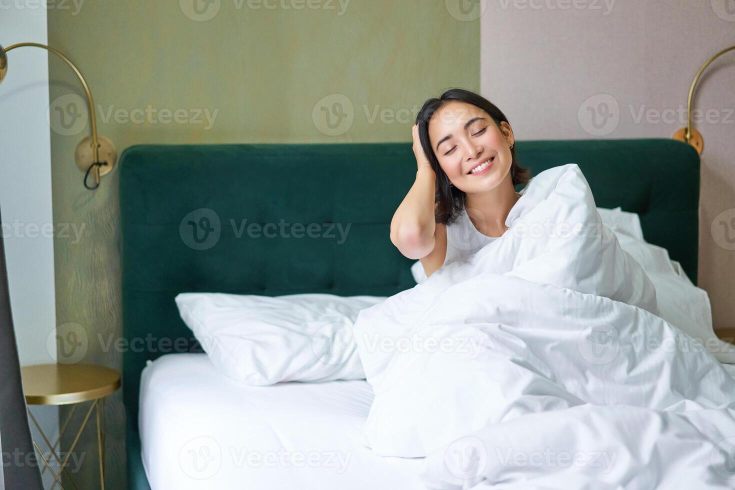 gelukkig Koreaans vrouw wordt wakker omhoog in haar hotel kamer, aan het liegen in bed onder knus warm dekens, wit lakens, genieten van weekend ochtend- foto