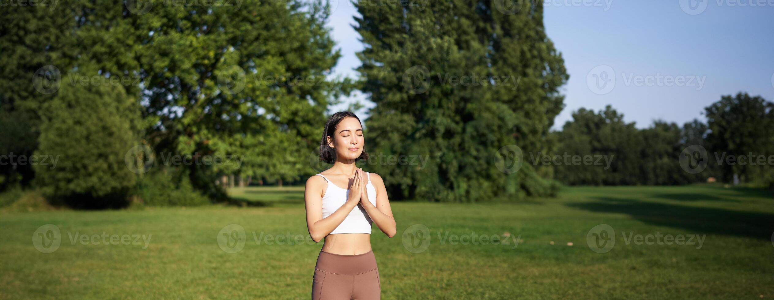 verticaal schot van Aziatisch vrouw staand in asana, aan het doen yoga opdrachten Aan vers lucht in park, vervelend leggings, staand Aan rubber mat foto