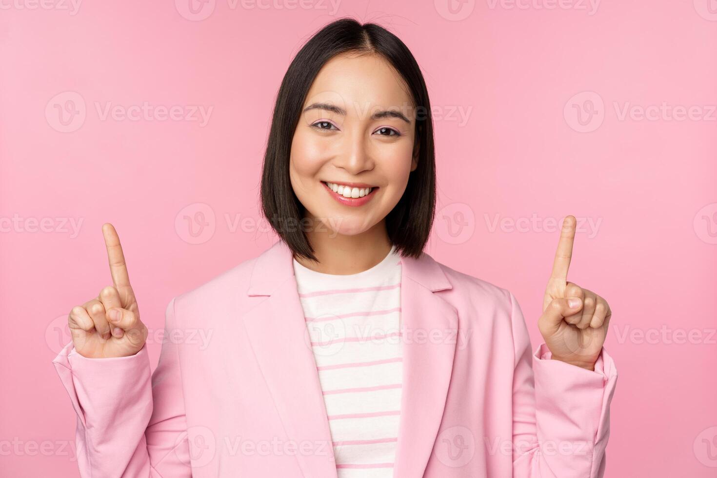 portret van Aziatisch zakenvrouw richten vingers omhoog en lachend, tonen bedrijf bedrijf logo, informatie Aan bovenkant, staand over- roze achtergrond foto