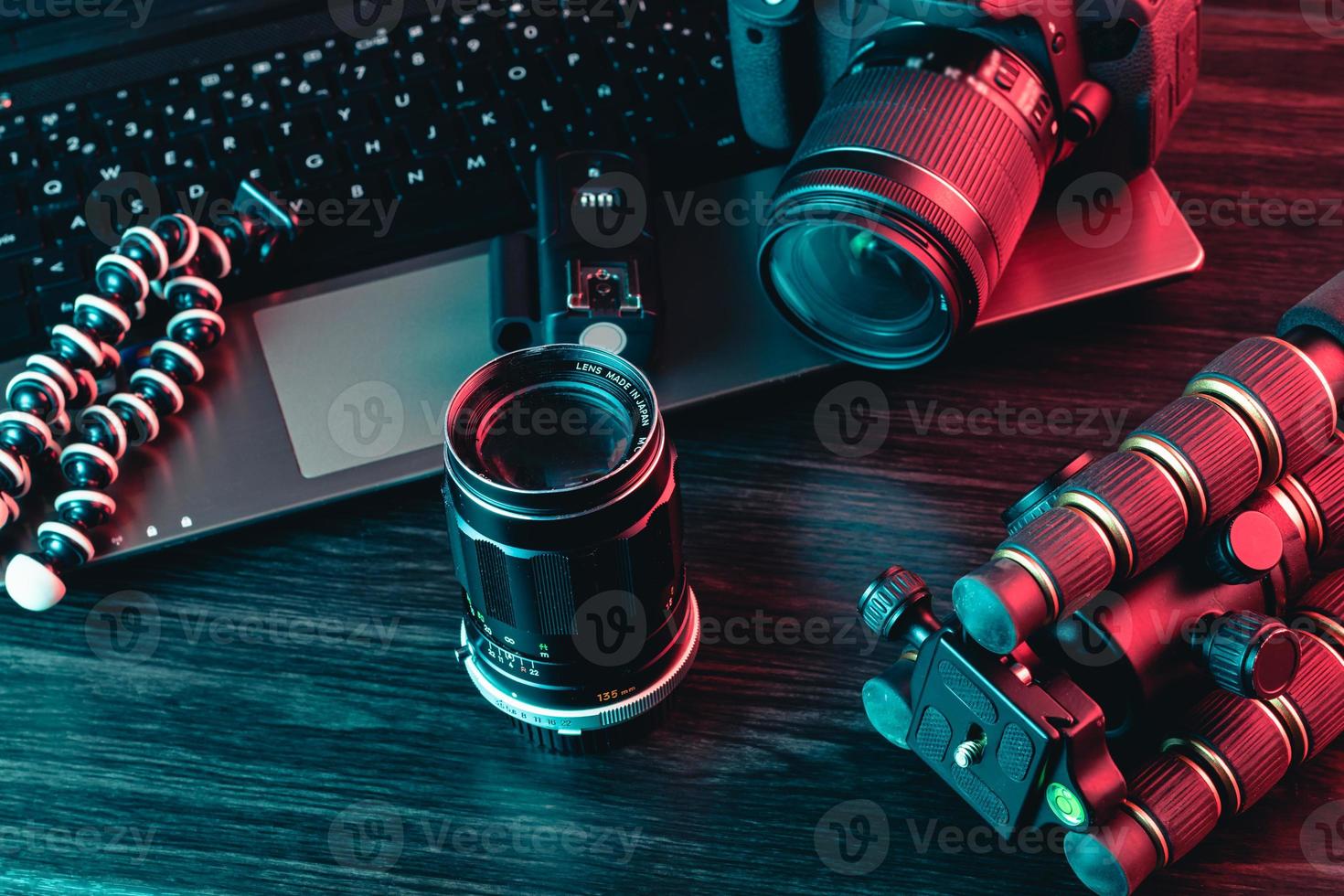 een werkruimte met laptop, moderne camera, lens, statief en een zwarte pen op een houten ondergrond. freelance of creatieve man of vrouw. rood en blauw licht foto