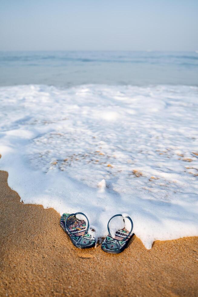 blauw strand slippers Aan een zanderig strand. de Golf wast de slippers uit de oever. rust uit door de zee, zomer avonturen. surfen lawaai en zee schuim foto