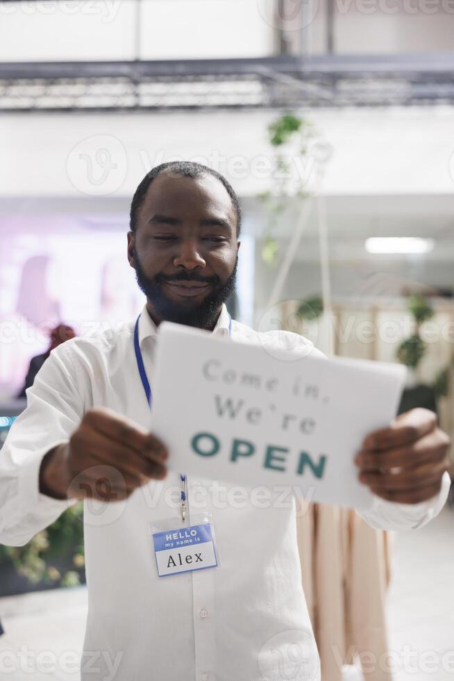 mode winkel gelukkig Afrikaanse Amerikaans Mens assistent hangende opening teken Aan winkel venster. glimlachen vrolijk kleding op te slaan werknemer Holding teken bord in de gaten hebben kopers over werk dag begin foto