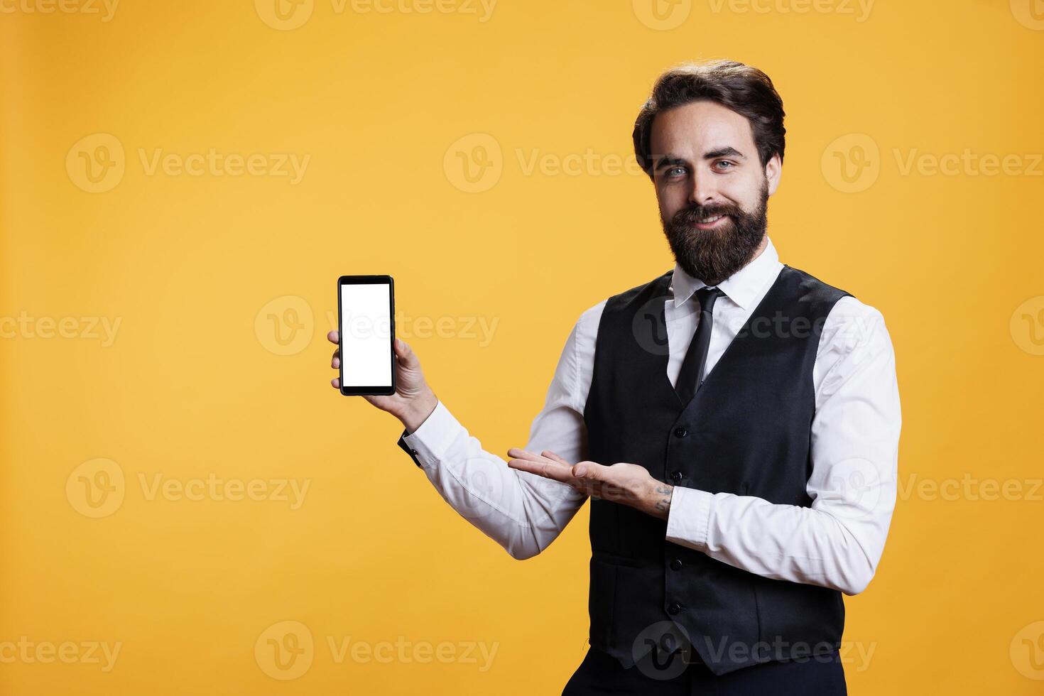 mannetje ober shows wit scherm in studio, presenteren geïsoleerd copyspace sjabloon Aan smartphone Scherm. professioneel elegant butler Holding leeg mockup scherm over- geel achtergrond. foto