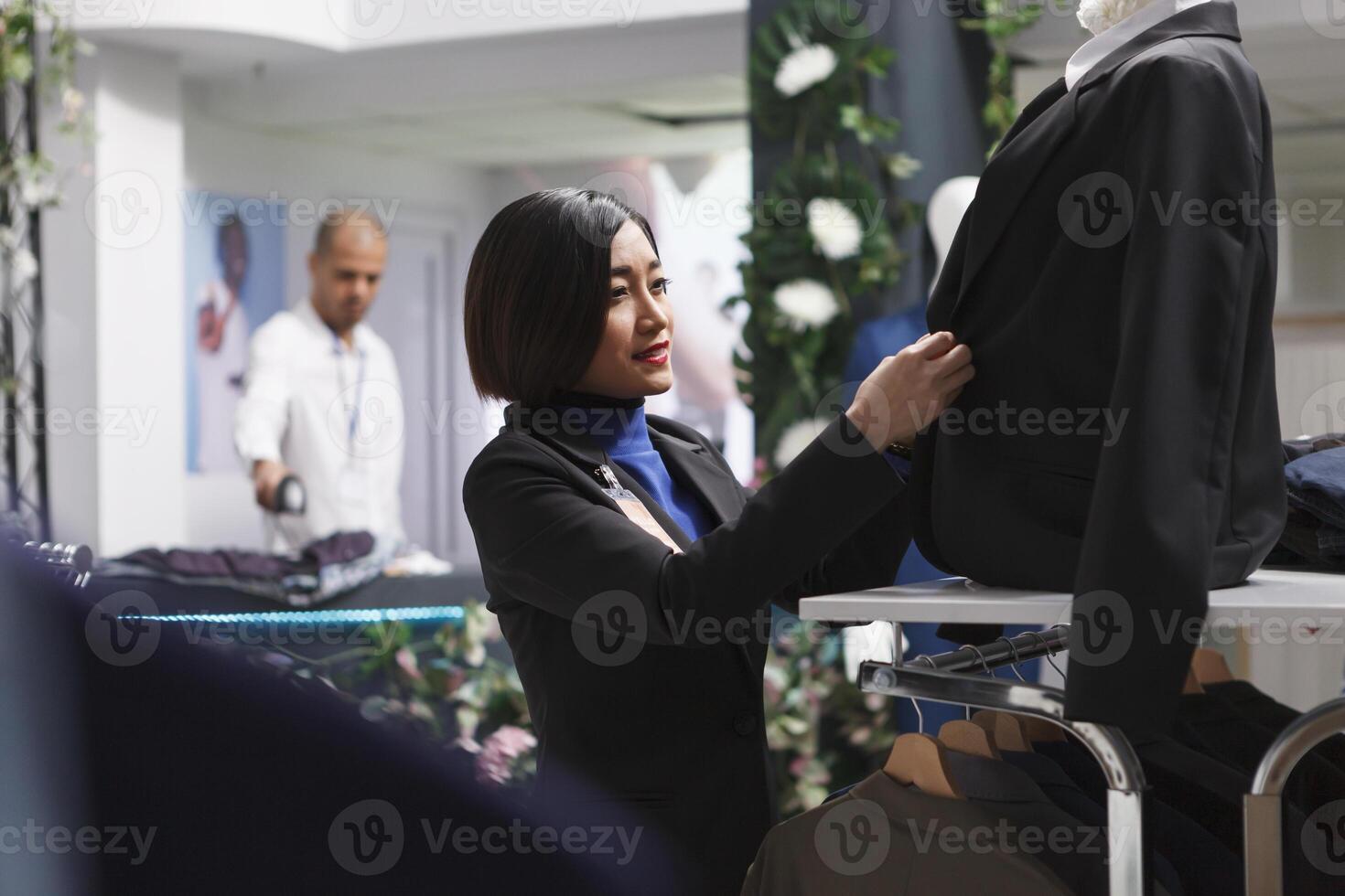 winkel Aziatisch vrouw arbeider vervelend mannequin in formeel vrouw jasje terwijl organiserende handelswaar in kleinhandel centrum. kleding op te slaan verkoper bevestiging pak toetsen Aan dummy model- foto