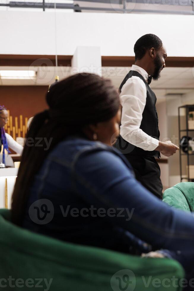 Afrikaanse Amerikaans hotel personeel en gast in lobby. vrouw reiziger aan het wachten voor Check in Bij ontvangst Oppervlakte, vriendelijk glimlachen loopjongen helpen gasten met bagage. gastvrijheid en toerisme industrie foto