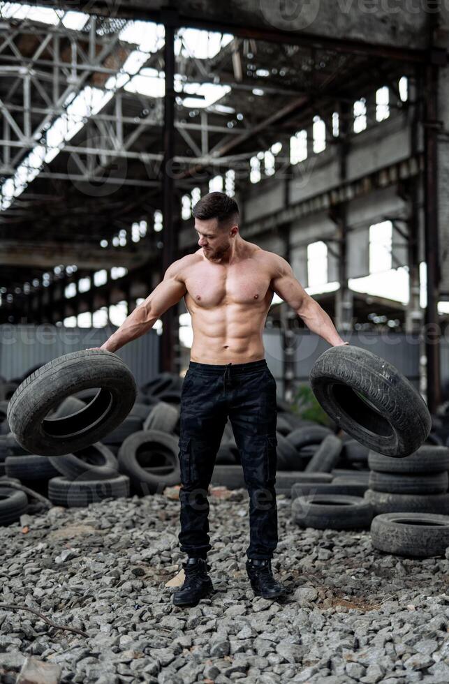 sportief Mens Holding zwaar banden. knap Mens met naakt buikspieren in jeans. verlaten huis Aan achtergrond. foto