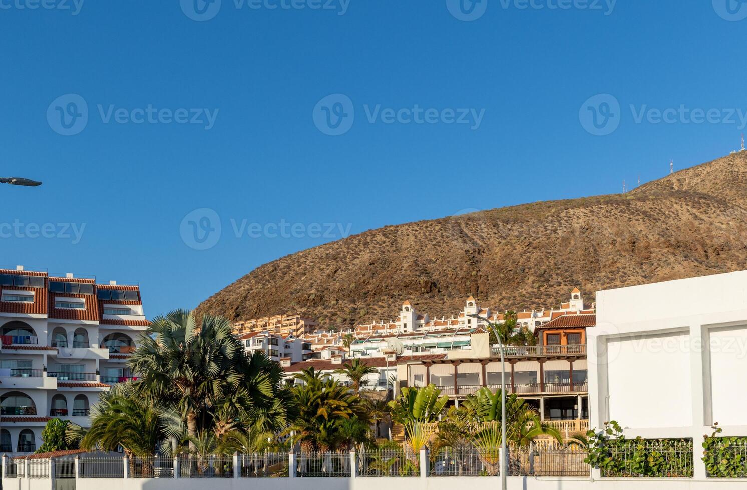 toneel- visie van een kust- stad- met wit gebouwen en geparkeerd auto's, reeks tegen een backdrop van bergen onder een Doorzichtig blauw lucht in los Cristianos, tenerife. foto