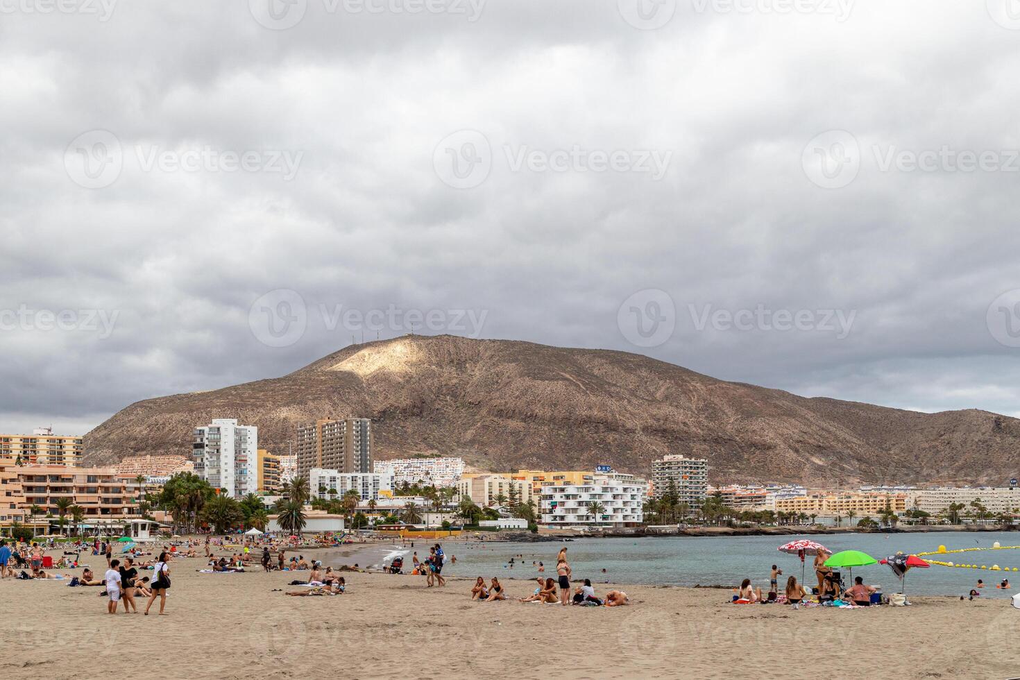 toneel- visie van een strand met mensen ontspannend, stadsgezicht, en een groot berg onder bewolkt luchten in los Cristianos, tenerife. foto