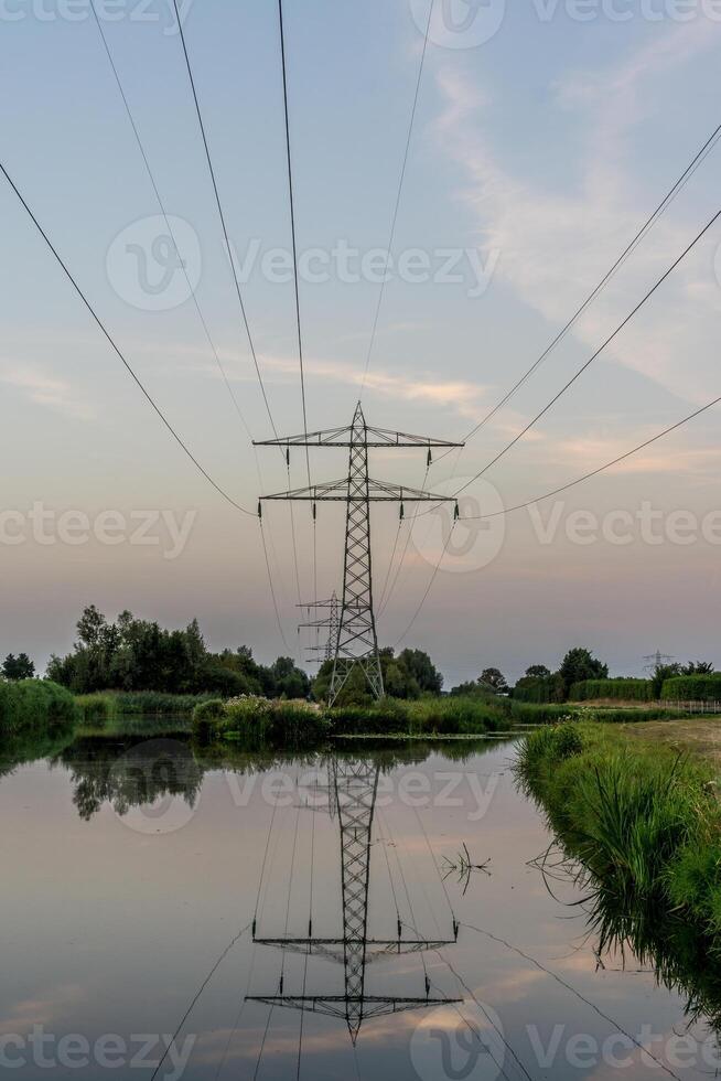 hoog Spanning kabels met pylonen wezen weerspiegeld in een vijver gedurende zonsondergang met een blauw lucht foto