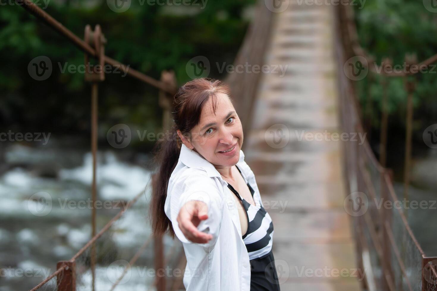 een volwassen vrouw strekt zich uit haar hand- naar jij, uitnodigend u naar wandelen aan de overkant de suspensie brug. foto