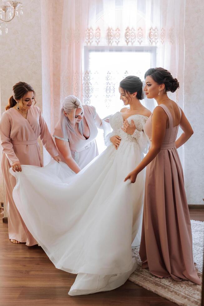 bruiloft ochtend. bruidsmeisjes helpen zetten Aan de wit bruiloft jurk. een jong vrouw is voorbereidingen treffen naar ontmoeten haar bruidegom en hebben pret met haar vrienden foto