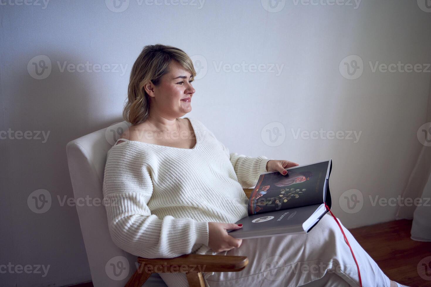 een middelgroot vrouw in licht kleren leest een boek terwijl zittend in een wit stoel in een licht kamer foto