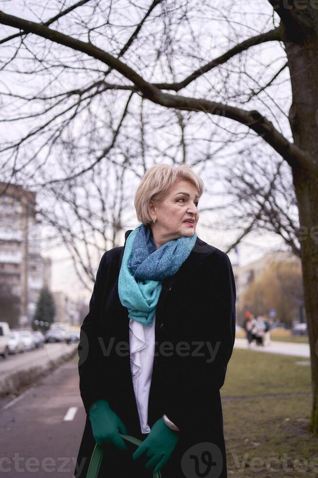 portret van een elegant oud vrouw in een zwart jas met groen accessoires Aan een voorjaar straat foto