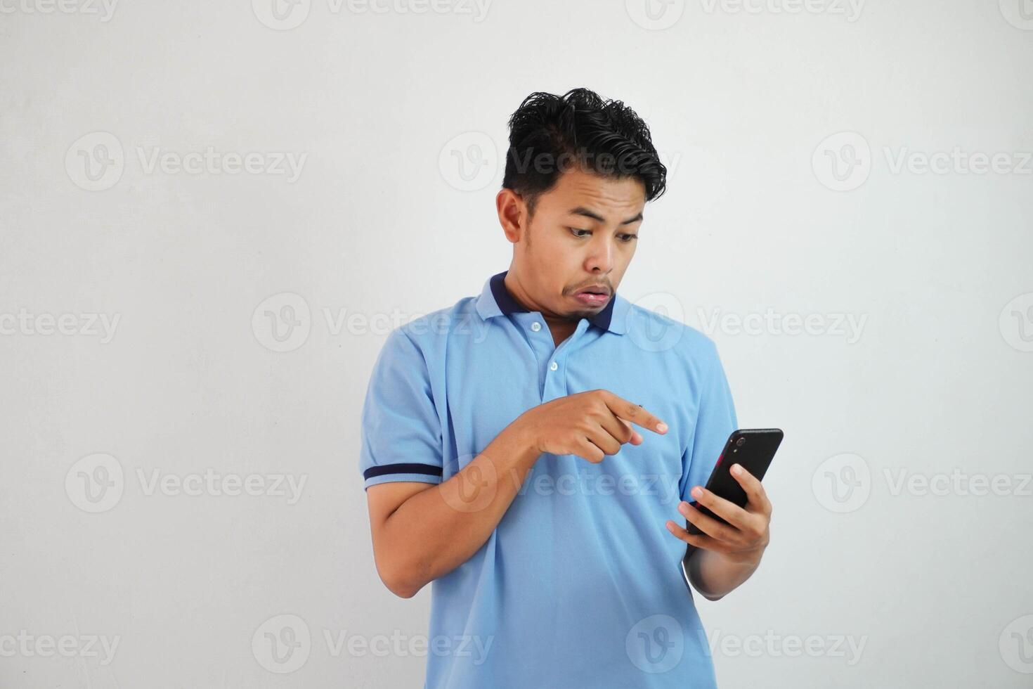 portret Aziatisch Mens geschokt Holding telefoon en richten Bij de telefoon met een vinger vervelend blauw polo t overhemd geïsoleerd Aan wit achtergrond foto