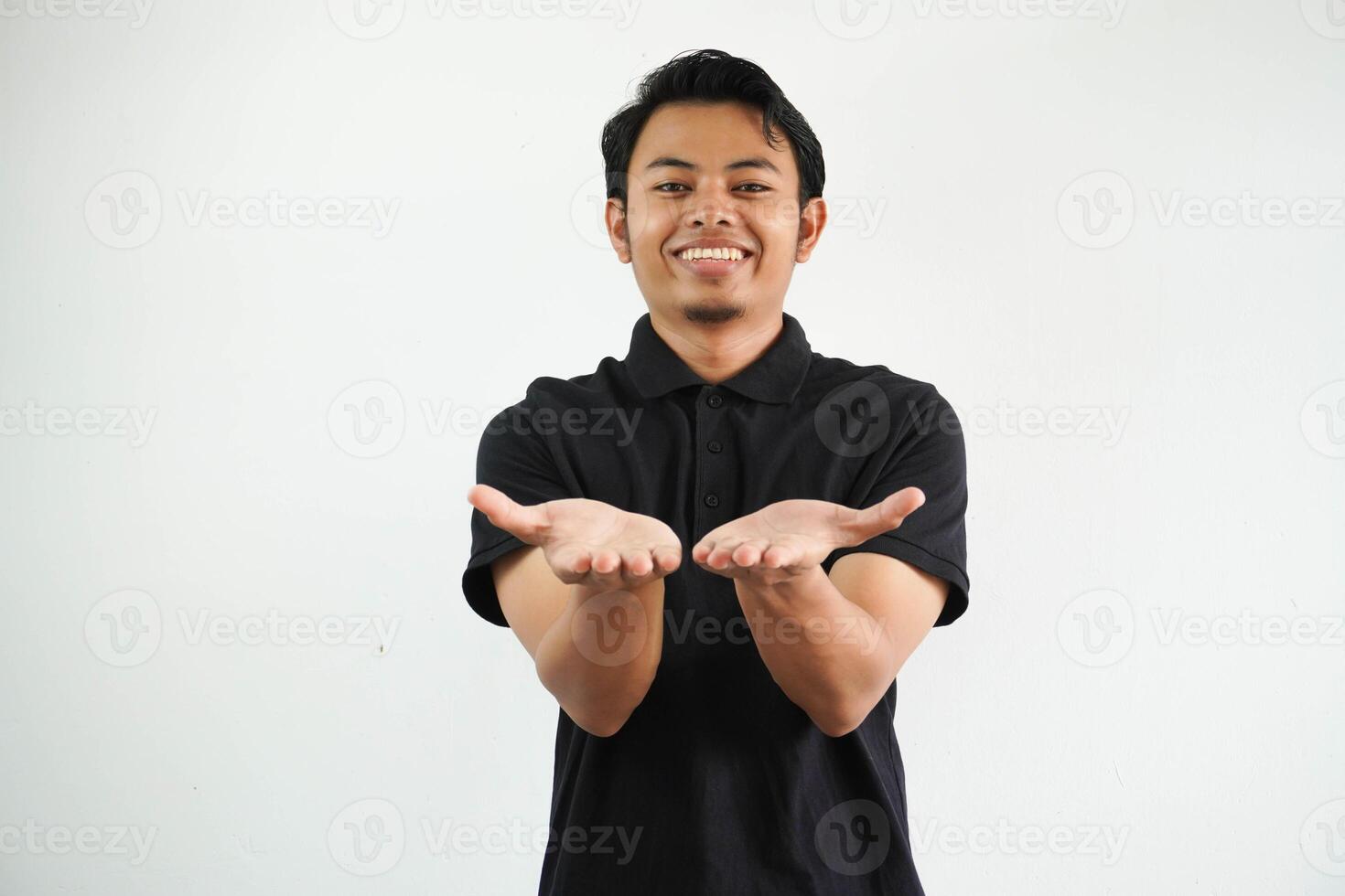 glimlachen jong Aziatisch Mens poseren Aan een wit backdrop Holding iets met handpalmen, aanbieden naar camera, vervelend zwart polo t shirt. foto