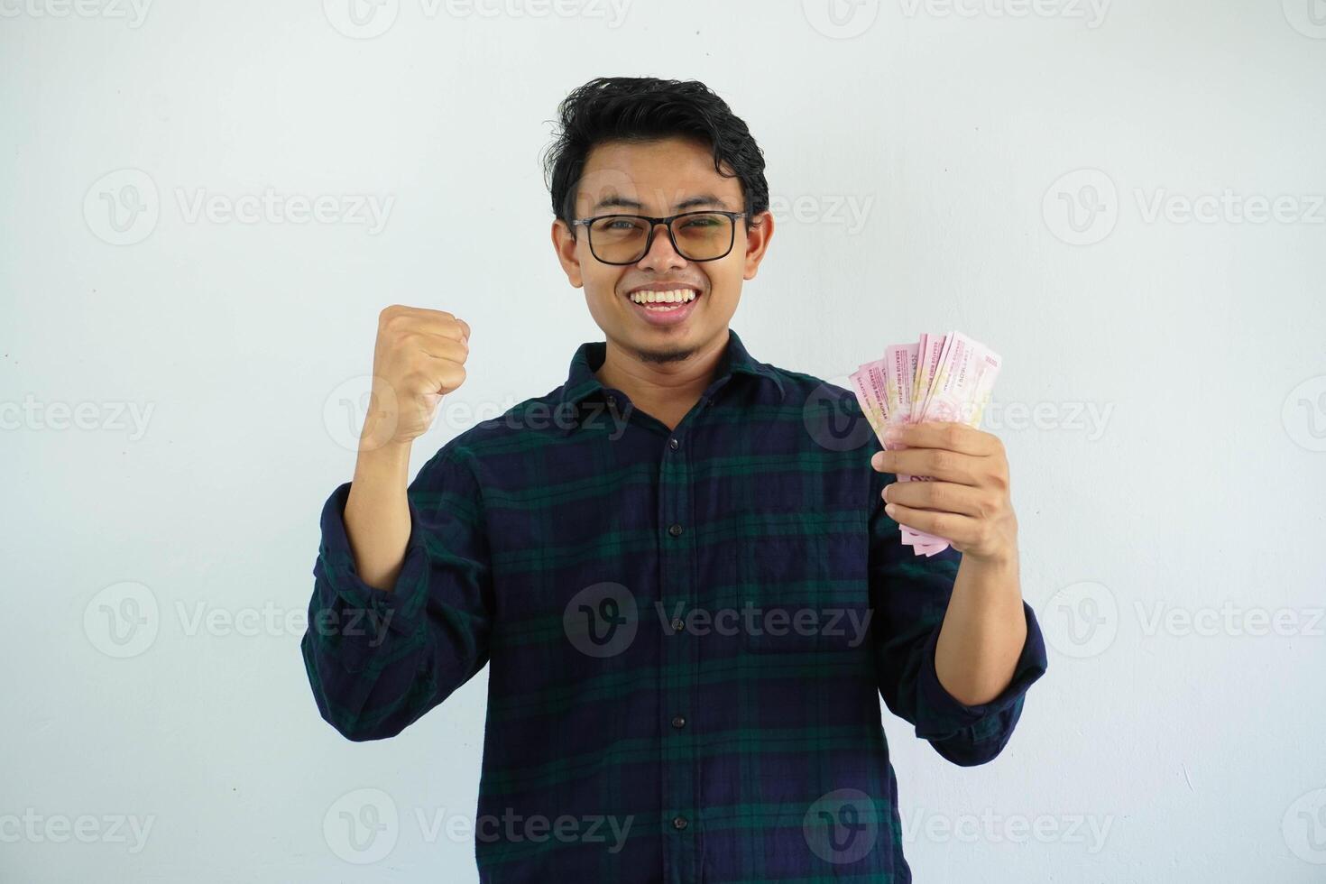 jong Aziatisch Mens gebalde vuist terwijl Holding Indonesië papier geld en tonen opgewonden uitdrukking geïsoleerd Aan wit achtergrond foto