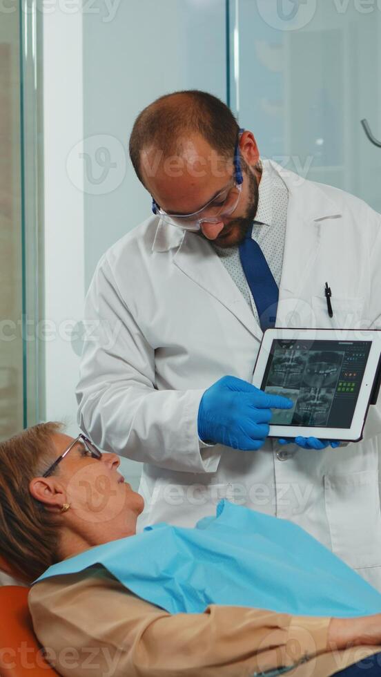 orthodontist gebruik makend van tablet uitleggen tandheelkundig X straal naar geduldig zittend Aan tandheelkundig stoel in stomatologisch kantoor. tandarts tonen naar oud vrouw radiografie gebruik makend van digitaal apparaat werken in modern kliniek. foto