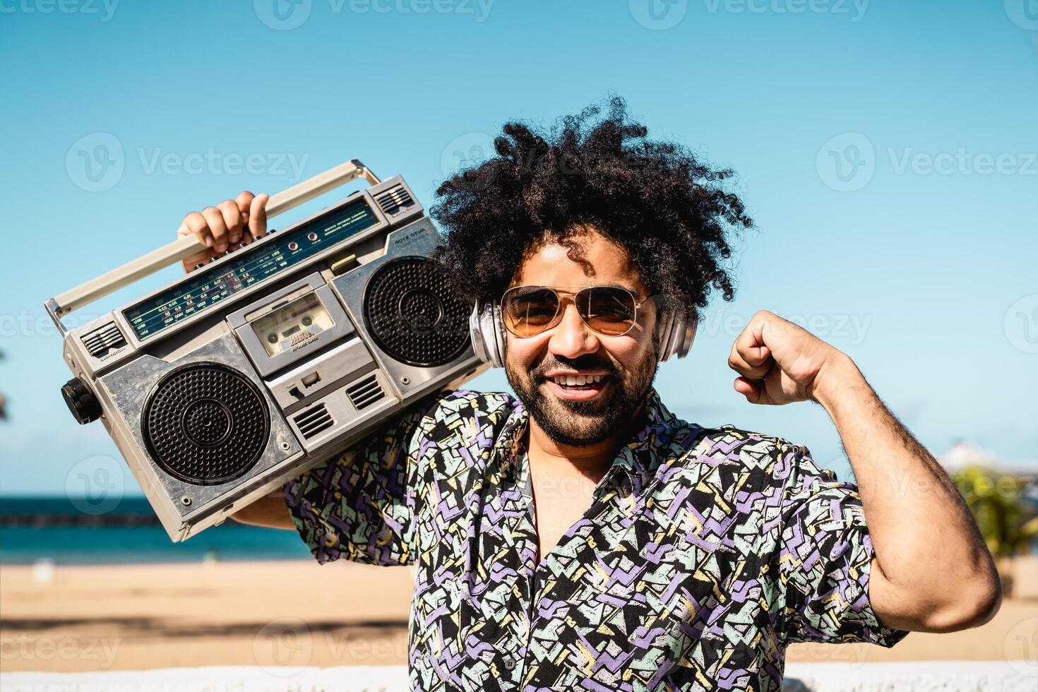 gelukkig Latijns Mens hebben pret luisteren naar muziek- met draadloze hoofdtelefoons en wijnoogst boombox Aan de strand gedurende zomer tijd foto