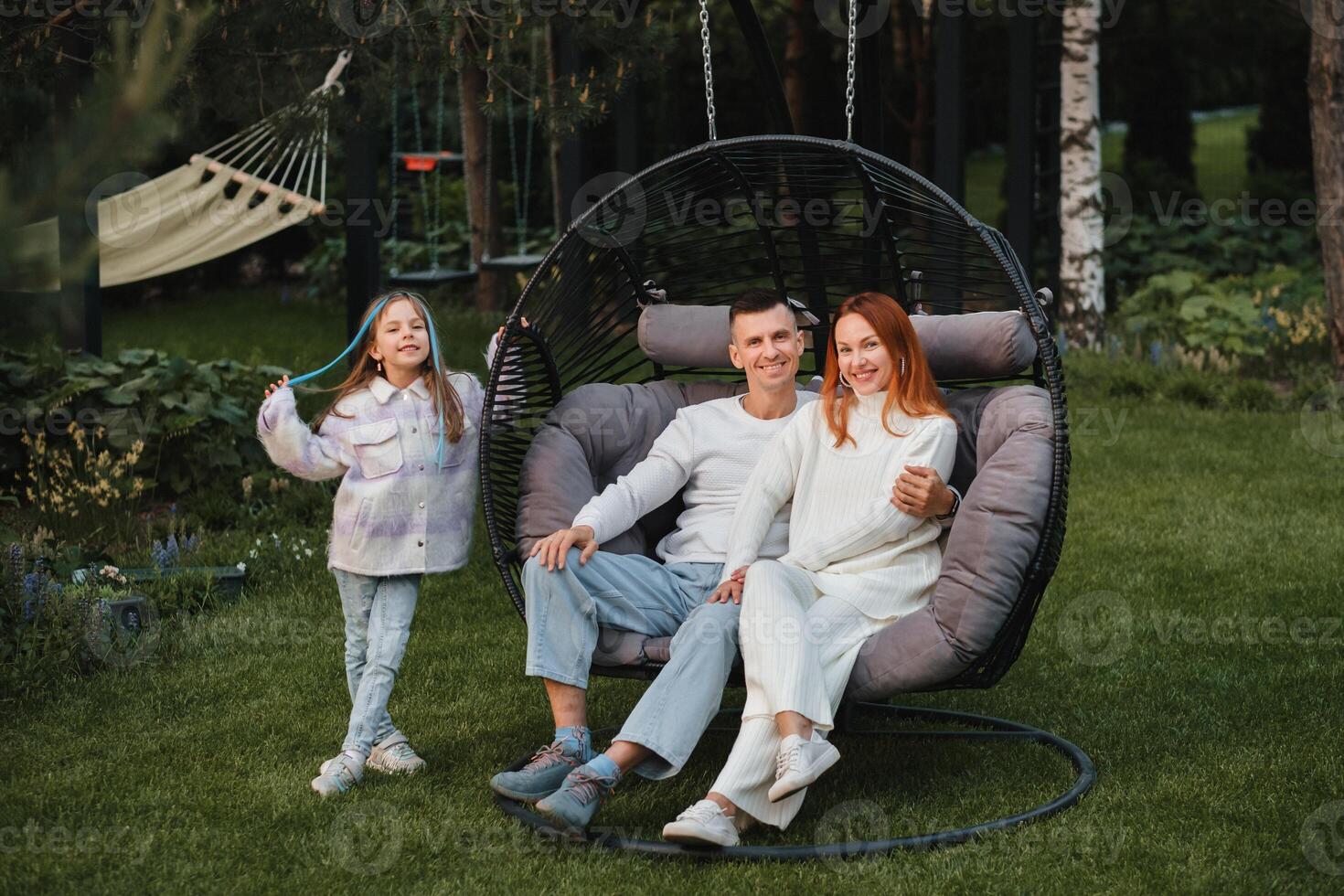 een gelukkig familie is zittend in een hangmat Aan de gazon in de buurt de huis foto