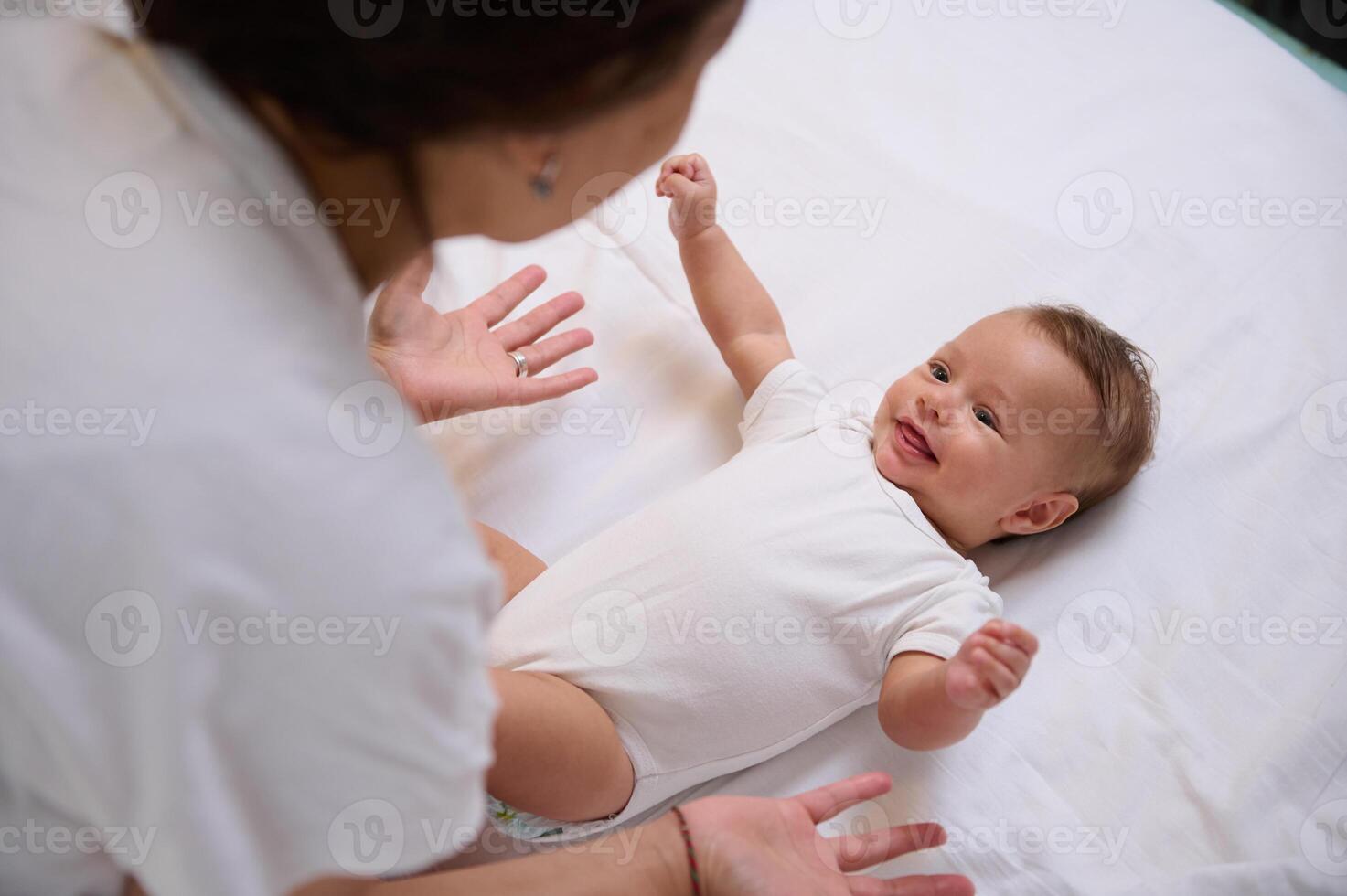 aanbiddelijk Kaukasisch baby jongen 4 maanden oud aan het liegen Aan de bed, houden zijn armen uitgestrekt, glimlachen naar zijn moeder foto