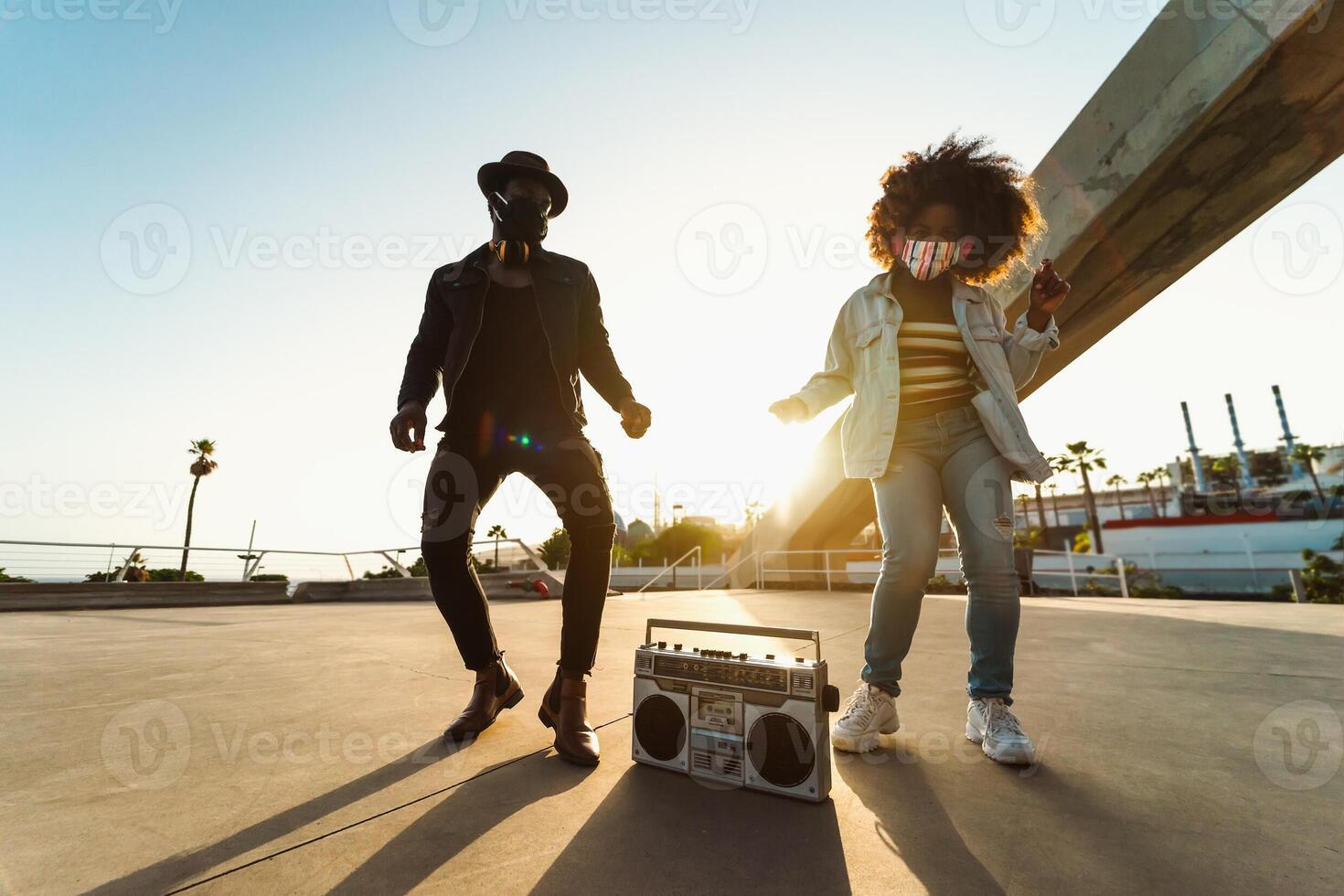 jong afro vrienden vervelend gezicht masker dansen buitenshuis terwijl luisteren naar muziek- met draadloze hoofdtelefoons en wijnoogst boombox foto