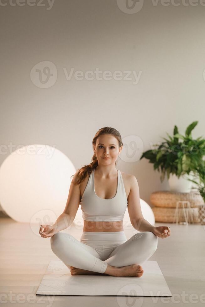 portret van een meisje in wit kleren zittend in een lotus positie Aan een mat Bij een binnen- yoga klasse foto