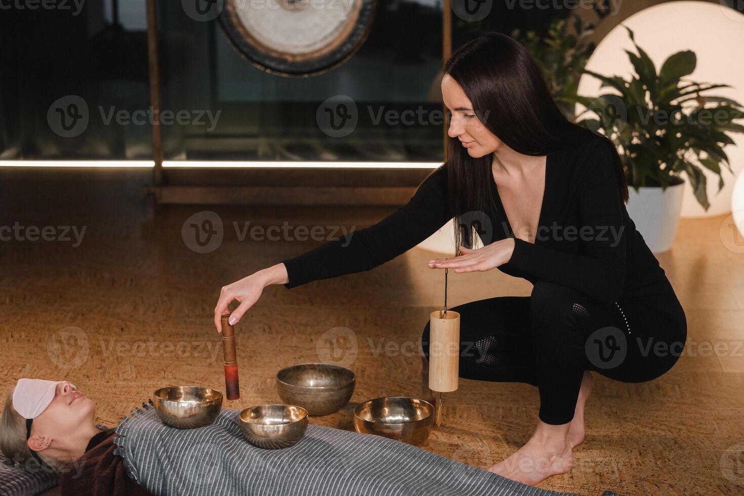 een jong mooi vrouw is aan het doen een massage met het zingen kommen en een koshi klok naar een ander meisje. geluid behandeling foto