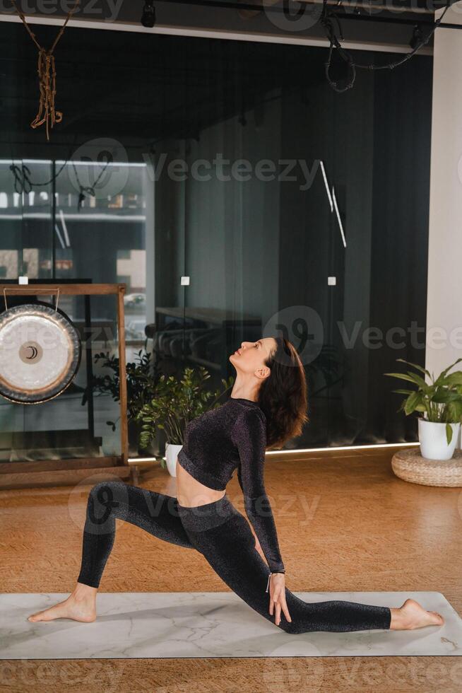 een meisje in een zwart sport- uniform doet yoga staand Aan een mat in de Sportschool foto