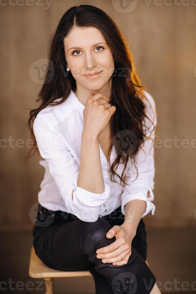 portret van een gelukkig jong brunette vrouw in een wit overhemd zittend Aan een stoel foto