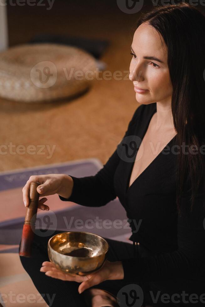 een vrouw in de lotus positie gebruik makend van een het zingen kom binnenshuis . ontspanning en meditatie. geluid therapie, alternatief geneesmiddel. boeddhistisch genezing praktijken foto