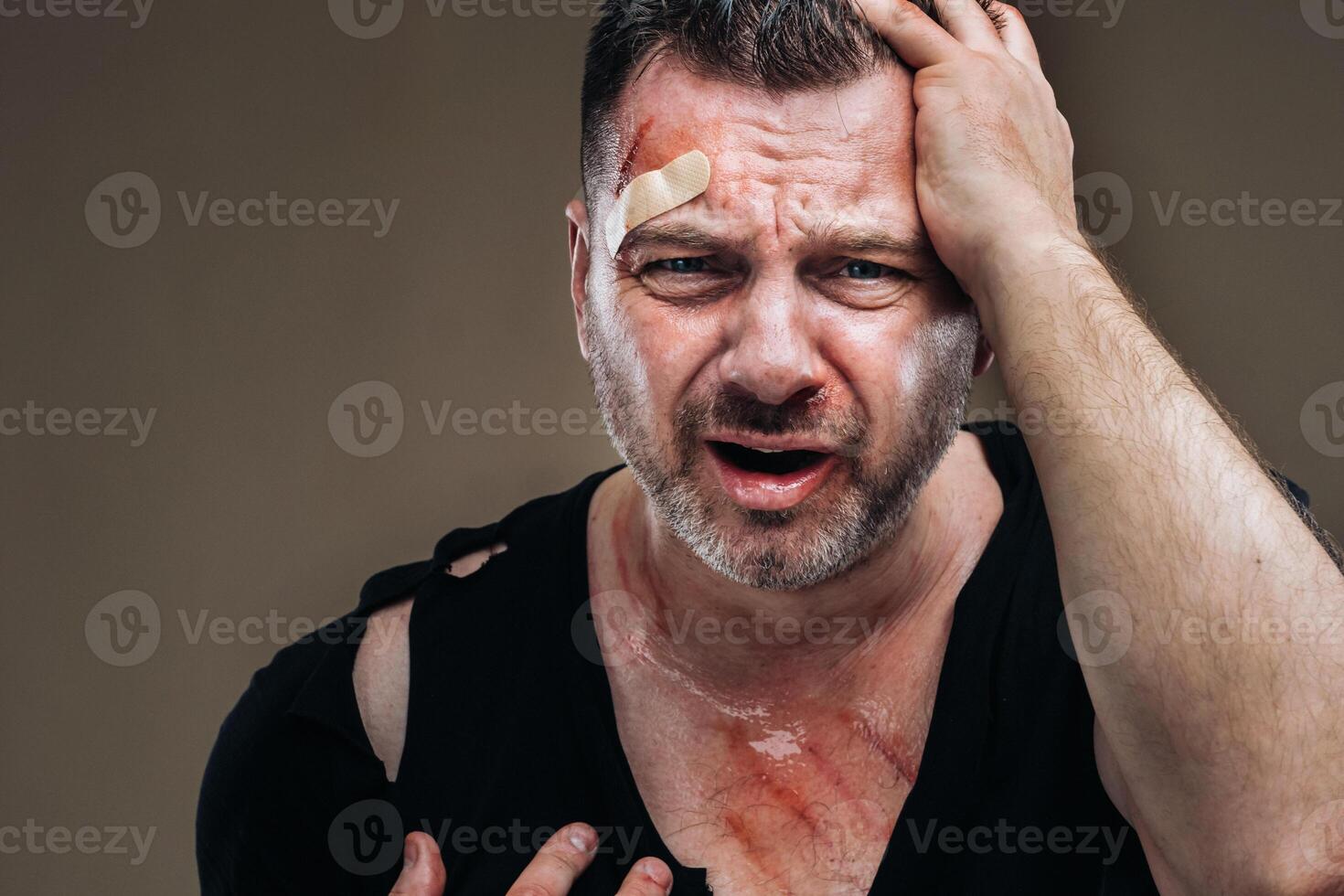 tegen een grijs achtergrond staat een gehavend boos Mens in een zwart t-shirt met wonden foto