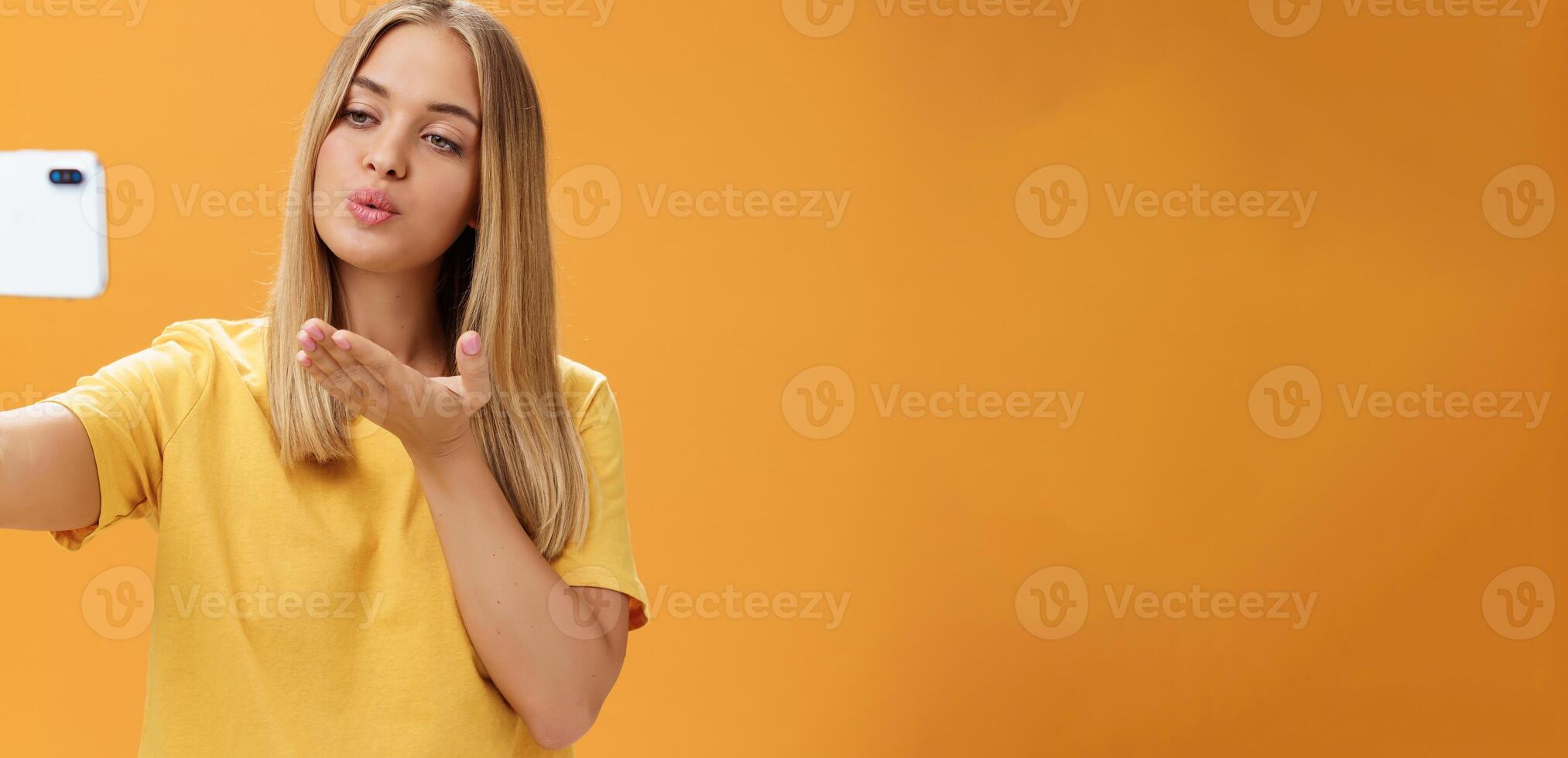 elegant betoverend vrouw mode blogger einde opname video via smartphone door Bezig met verzenden lucht kus Bij camera, nemen selfie met sensueel en zelfverzekerd blik Bij scherm poseren over- oranje muur foto