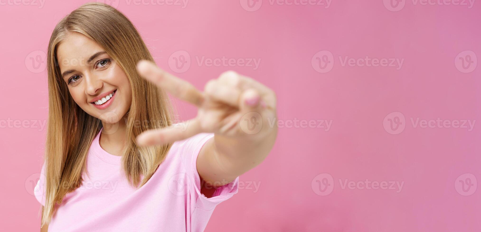 vredig en blij mooi jong meisje met Rechtdoor blond haar- in t-shirt trekken hand- met vrede teken naar camera, kantelen hoofd glimlachen vriendelijk poseren tegen roze achtergrond foto